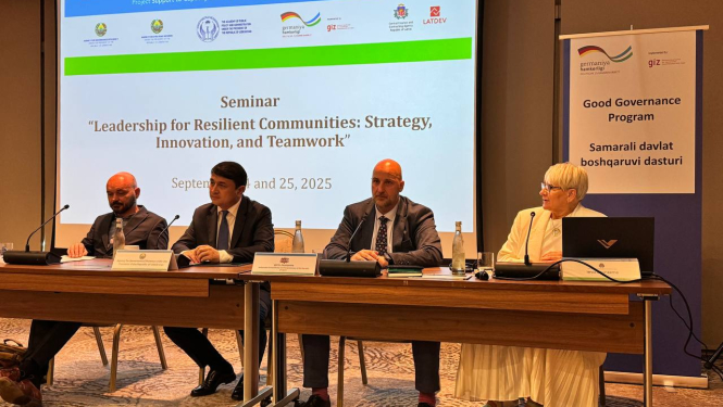 Seminārs “Līderība noturīgām kopienām: stratēģija, inovācijas un komandas darbs”, kas notika 2025. gada 24. un 25. septembrī. Attēlā redzami četri cilvēki, kas sēž pie garas galda ar mikrofonu priekšā. Fonā redzams liels ekrāns ar semināra nosaukumu, datumu un organizāciju logotipiem. Labajā pusē novietots plakāts ar uzrakstu “Good Governance Program” angļu un citā valodā. Telpa ir konferenču zāle ar paklāju un profesionālu iekārtojumu